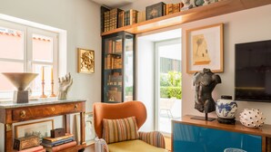 A cozy reading nook with a leather armchair, a wooden side table with books, a bookshelf, and a large window offering a view of the outdoors. - By the Castle | LUXURIOUS, PRIVATE GARDEN & HEATED SWIMMING POOL (Lisboa)