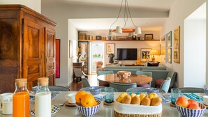 A modern living room with a dining area, featuring a wooden cabinet, a round table with breakfast items, and a cozy seating arrangement. - By the Castle | LUXURIOUS, PRIVATE GARDEN & HEATED SWIMMING POOL (Lisboa)