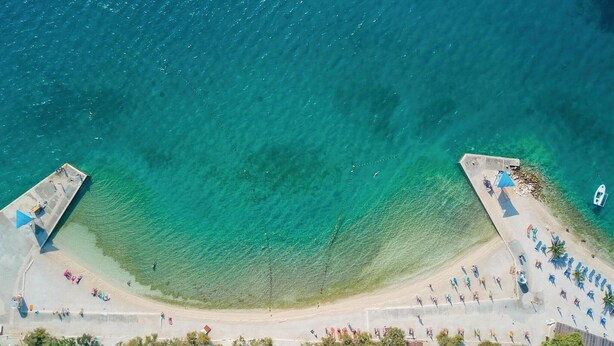 An aerial view of a beach with clear turquoise water, a pier on the left, and a sandy area with umbrellas and people on the right. - Nice apartment in Seget Donji with house sea view (Seget Donji)