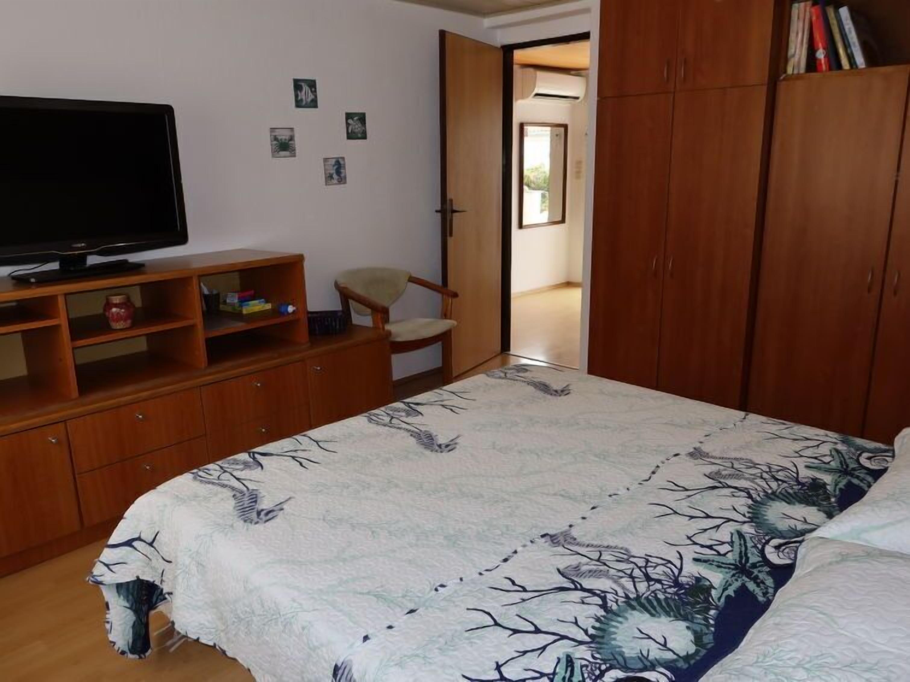A bedroom with a bed, a television, a wooden wardrobe, a chair, and a small shelf with books.