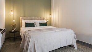 Un lit bien fait, recouvert d’une couvre-lit blanche et agrémenté de deux coussins verts, une table de chevet avec une horloge et un vase de fleurs séchées, et deux lampes suspendues au-dessus du lit.