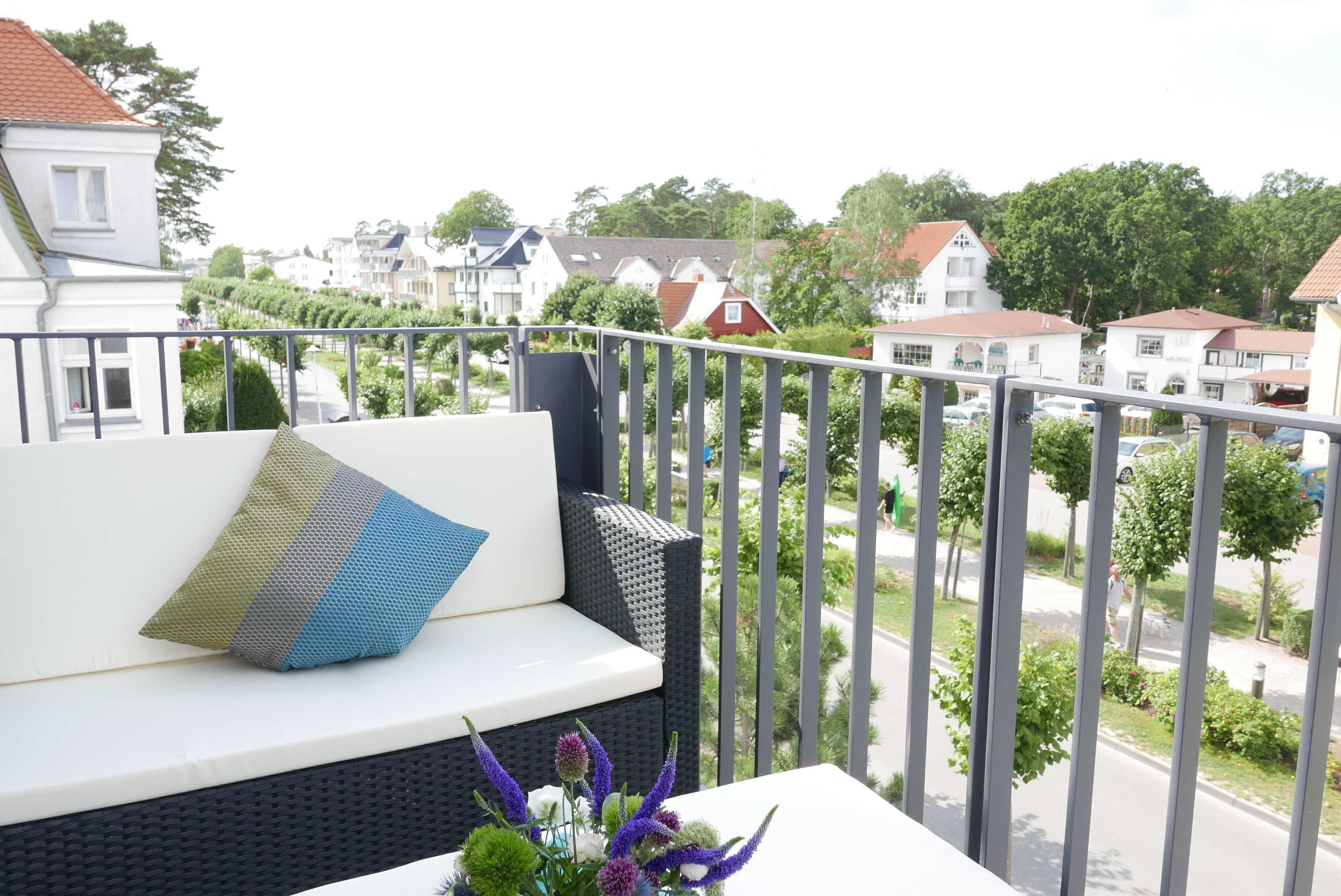 En balkon med en hvid sofa, gelænder af metal og udsigt over en boligvej med huse og træer.