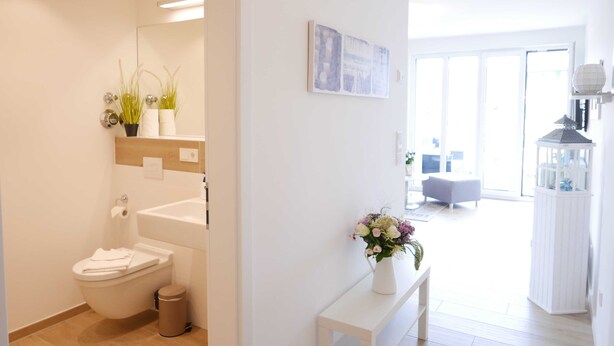 A modern bathroom with a toilet, sink, and mirror. There is a white bench with a vase of flowers in the living area.