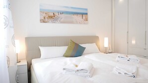 A neatly made bed with white linens and towels, a painting of a beach scene on the wall, and a large wardrobe.