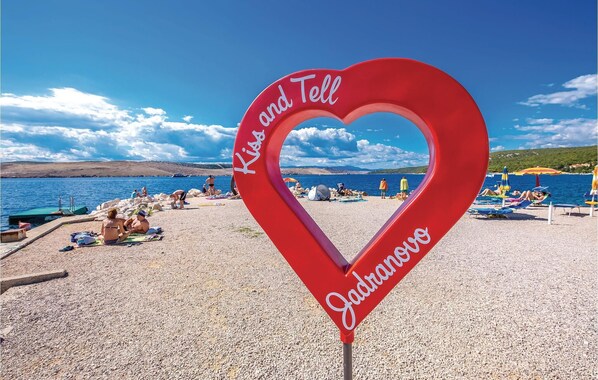 A heart-shaped sign with "Kids and Tell" and "Jadransko" written on it, placed on a pebbled beach with people and umbrellas in the background.