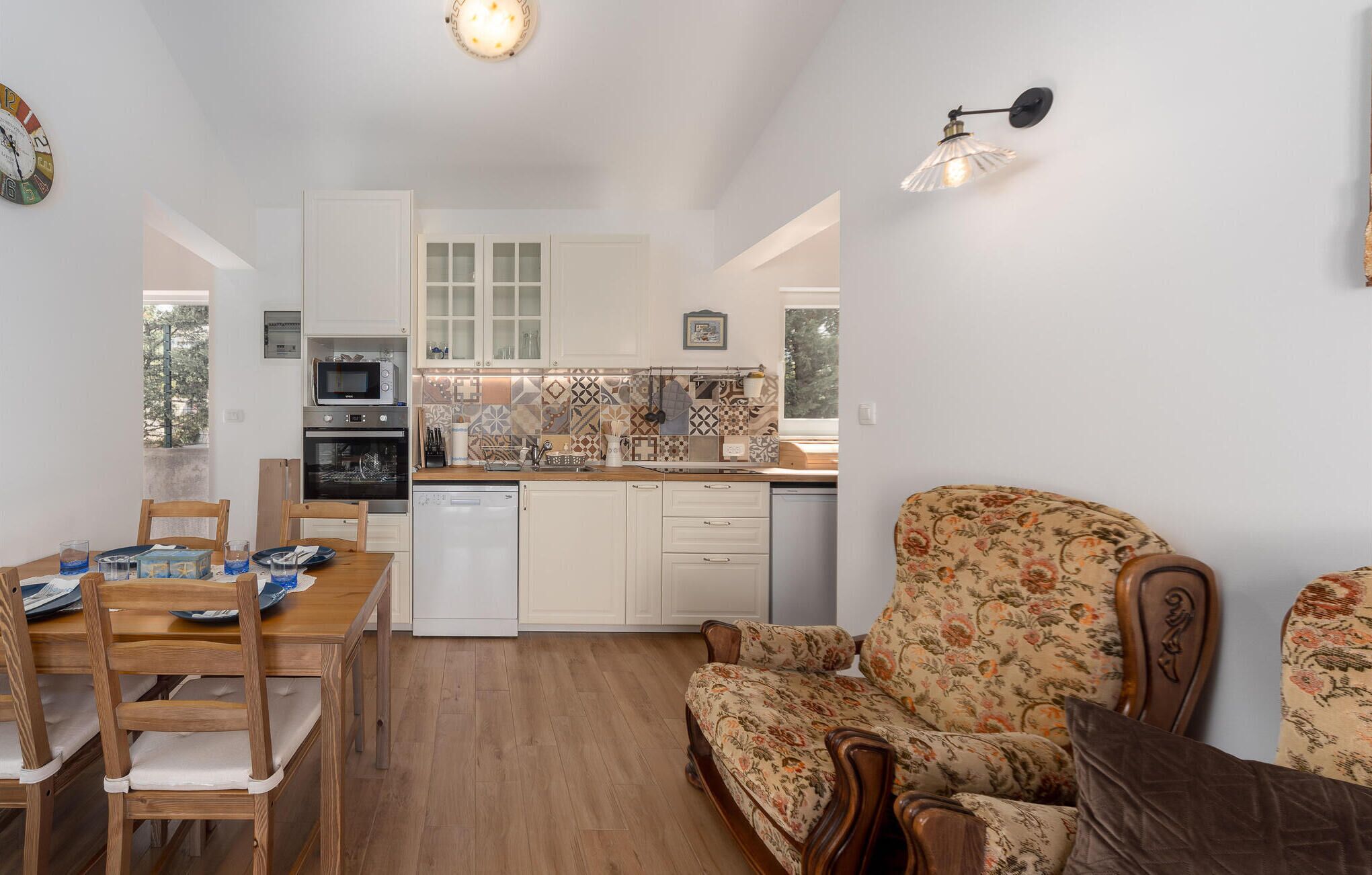 Eine kompakte Küche mit weißen Schränken, einer Mikrowelle, einem Backofen und einem Geschirrspüler. Es gibt einen Esstisch mit Stühlen und ein blumiges Sofa.