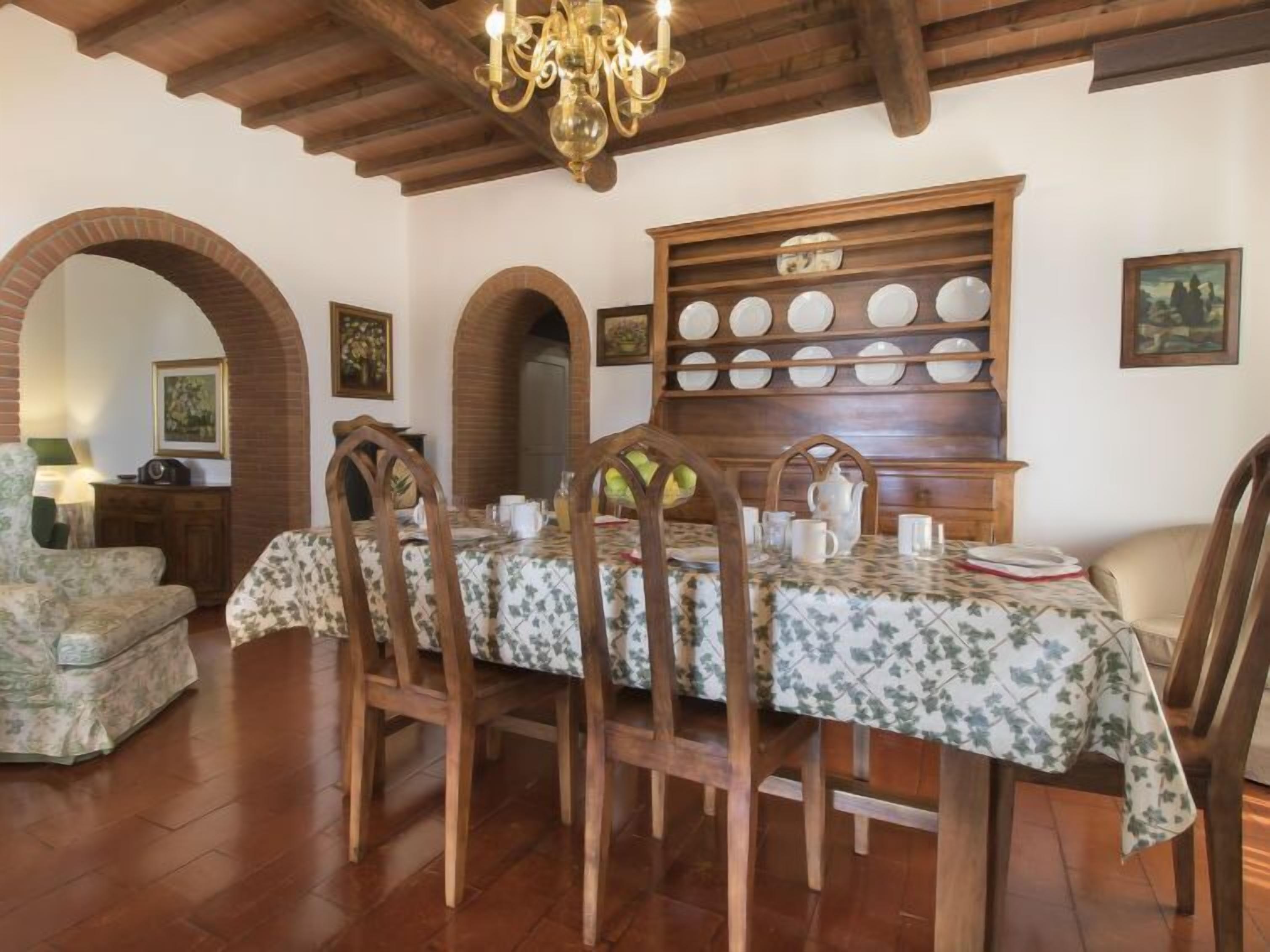 Un coin repas avec une table en bois dressée pour quatre personnes, entourée de chaises, d’un lustre et d’un porte-assiettes fixé au mur.