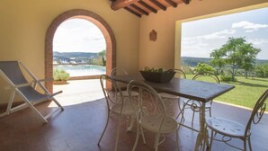 Un patio con mesa y sillas, vistas a una piscina y un paisaje con árboles y colinas.