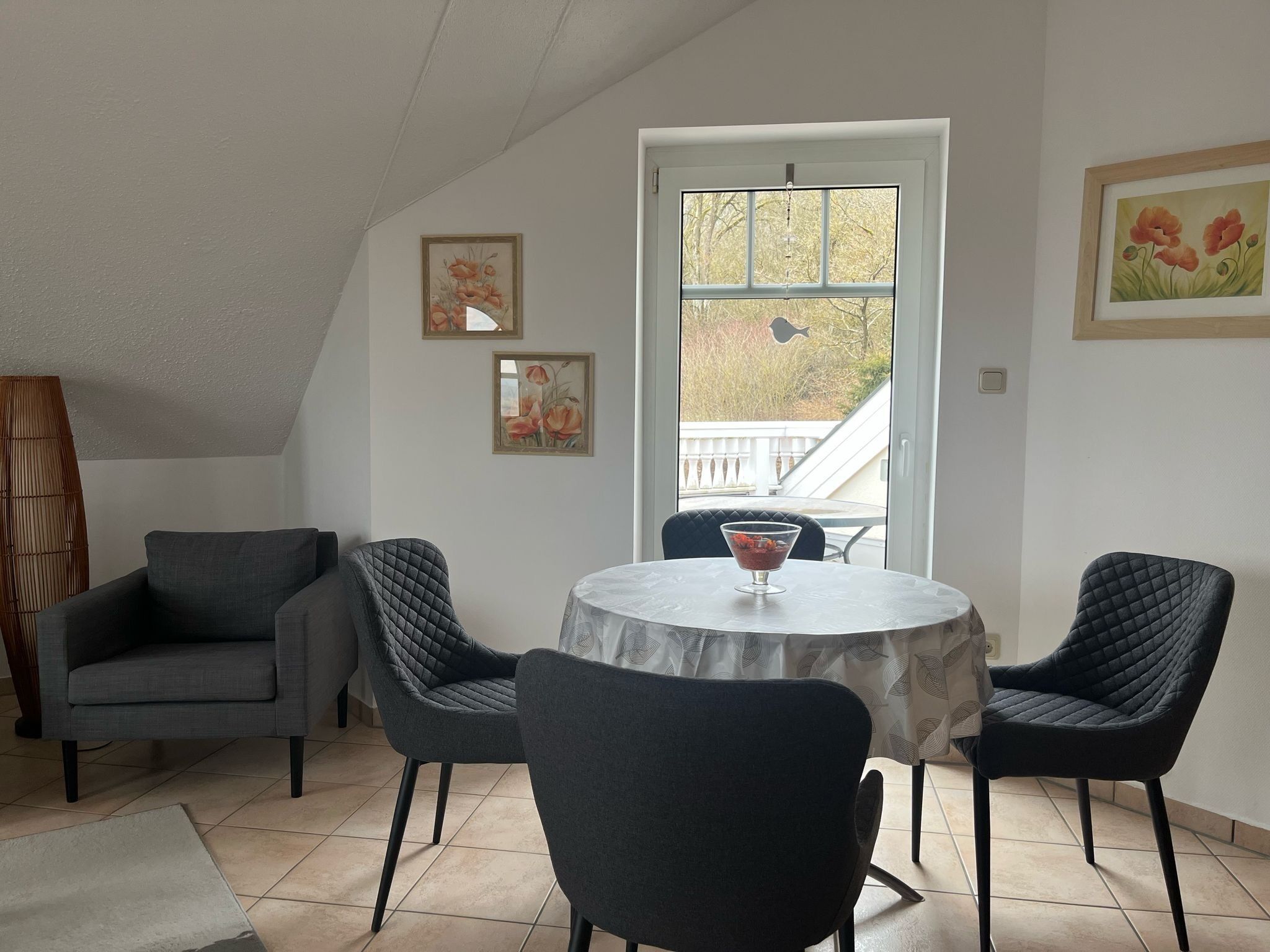 Un coin repas avec une table ronde, deux chaises, un vase de fleurs et une fenêtre donnant sur un balcon.