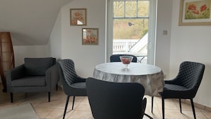A dining area with a round table, two chairs, a vase of flowers, and a window with a view of a balcony.