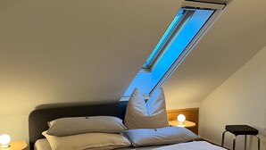 A bedroom with a skylight, a bed with pillows, a bedside table with a lamp, and a stool.