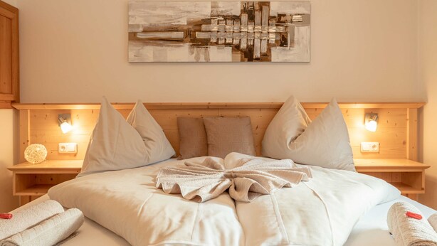 A hotel room with a large bed, wooden headboard, two bedside tables with lamps, and a wall-mounted abstract painting. - Apartment Dobratsch - Apartments Umfahrer Nassfeld (Tröpolach)