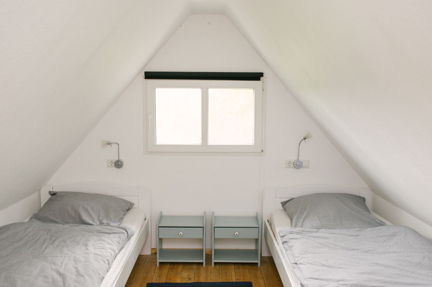 Une petite chambre mansardée triangulaire, avec deux lits simples, une fenêtre et une table de chevet placée entre les deux lits.