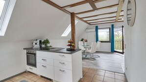 Ett modernt kök med vita skåp, en inbyggd ugn och en liten köksö med en kaffemaskin. Det finns en matplats med ett bord och stolar, och ett stort fönster med utsikt över omgivningen.