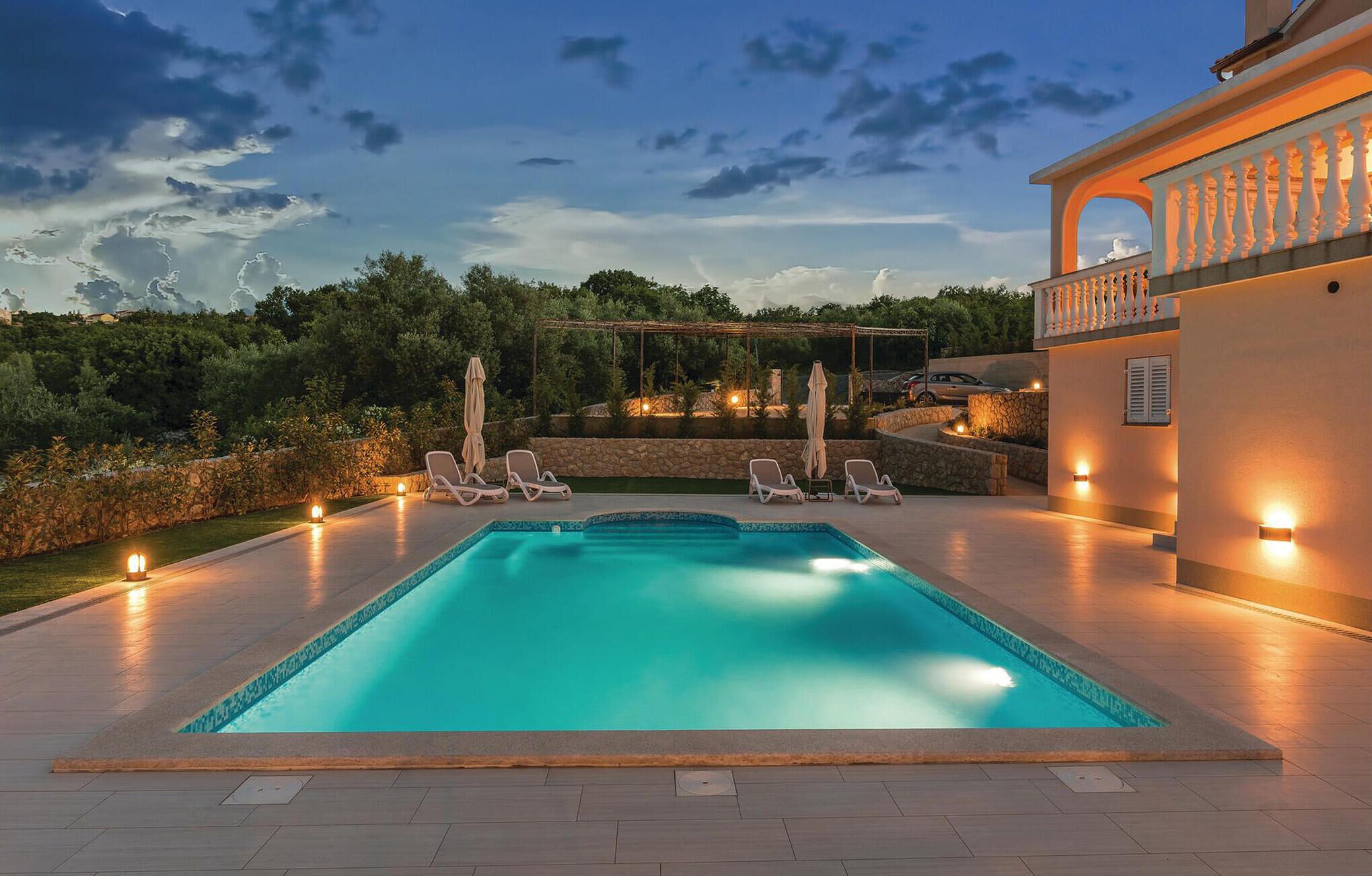 A rectangular swimming pool with a stone border, surrounded by lounge chairs and umbrellas. There is a stone wall and a pergola with lights.
