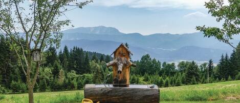 A wooden sculpture of a horse's head on a log, with a small yellow watering can beside it, set against a backdrop of mountains and trees.