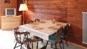 En spiseplads med et bord dækket til seks personer, omgivet af træstole, en lampe og et fjernsyn på et træskab.
