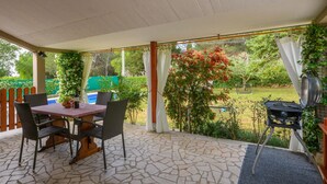 A covered patio with a stone floor, a table with chairs, a grill, and a view of a garden with trees and a pool. - Gorgeous home in Krnica (Krnica)