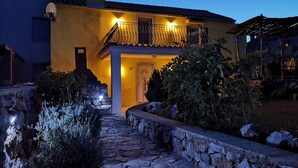 Una casa de dos pisos con un camino de piedra que conduce a la entrada, un balcón con barandilla y un pequeño jardín con arbustos y un muro de piedra.