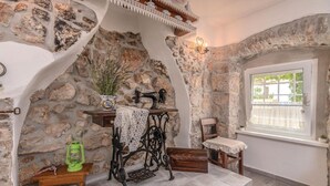 Una habitación rústica con pared de piedra, una máquina de coser antigua, una mesa de madera, un farol verde y una ventana con vistas.