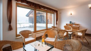 A room with wicker furniture, a table with glasses, a bottle, and a view of a building through large windows. - Double room - Ferienhof Stadlpoint (Ried im Zillertal)