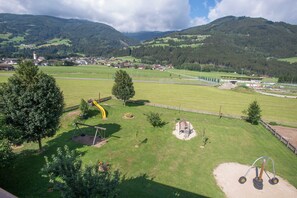 A playground with swings and a slide, surrounded by green fields and mountains in the background.