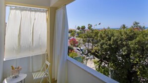 Un balcón con una silla y mesa blancos, una ventana con cortinas blancas y vista a una playa y edificios.