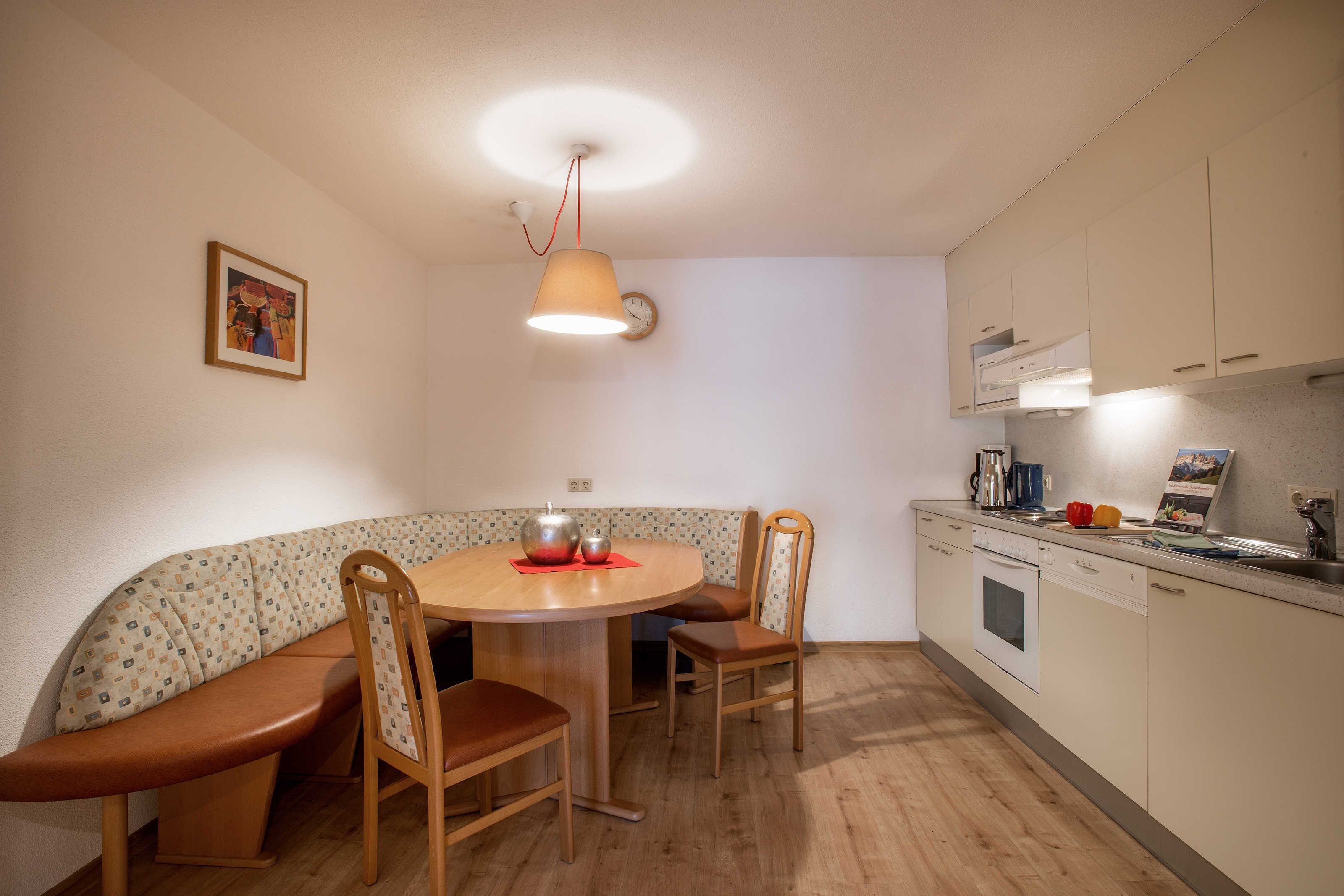 A compact kitchen with a round dining table, wooden chairs, a built-in oven, and a clock on the wall.
