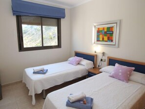 A room with two single beds, a window with blinds, a wall-mounted light, and a colorful painting.