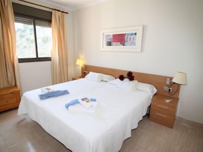 Un cuarto de hotel con una cama grande, dos mesitas de noche, una ventana con cortinas, un cuadro en la pared y una lámpara en la mesita.