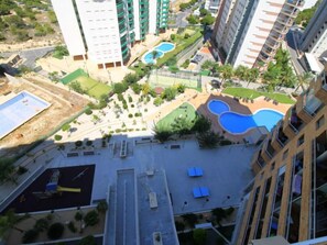 A high-rise building with multiple balconies, a rooftop pool, and a landscaped courtyard with a smaller pool and tennis court.