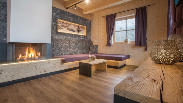 Una sala de estar acogedora con chimenea, un muro de acento de piedra, un banco de madera y un ventanal con cortinas.