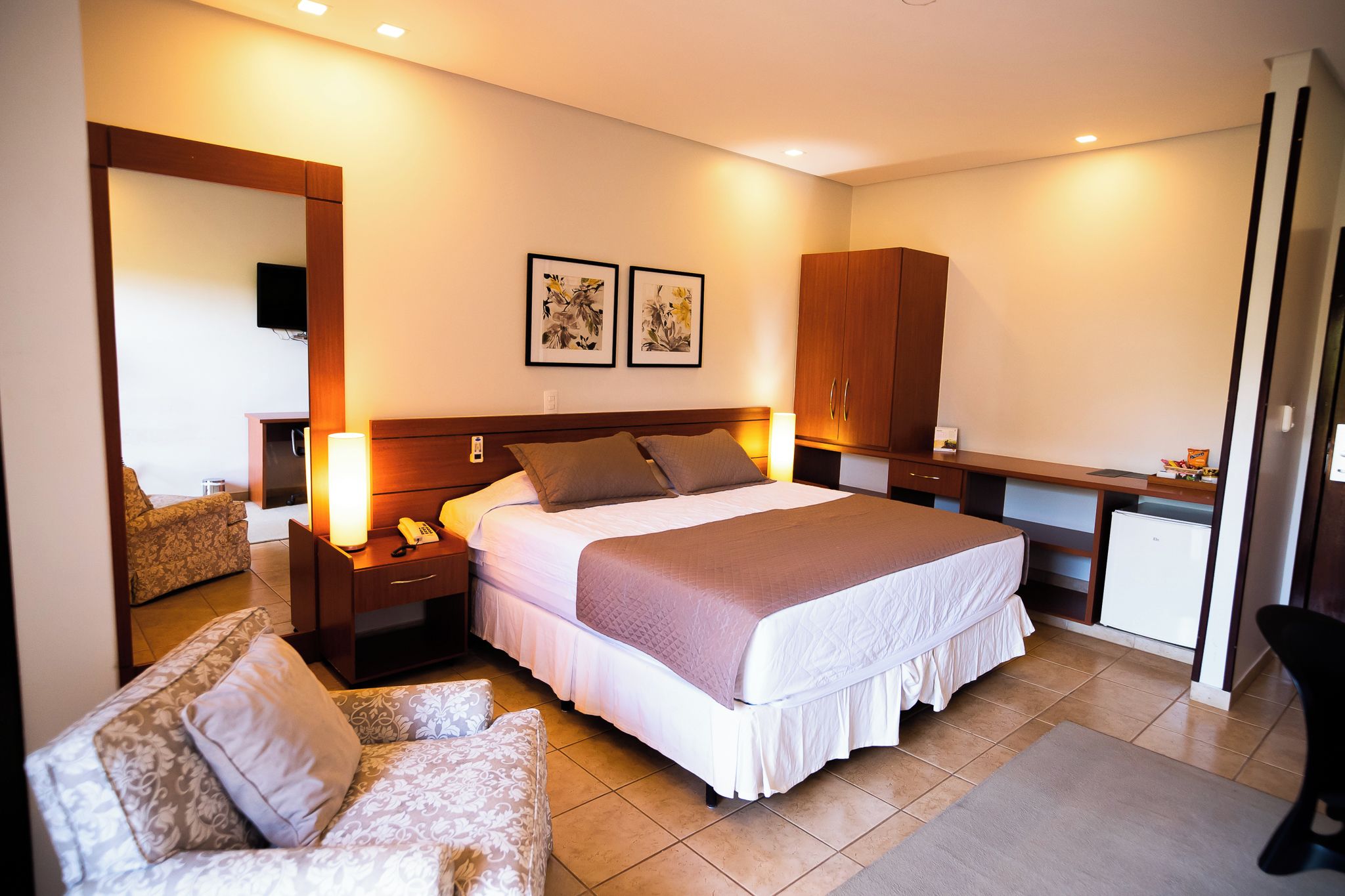 A hotel room with a bed, bedside tables, a desk, a chair, and artwork on the wall.