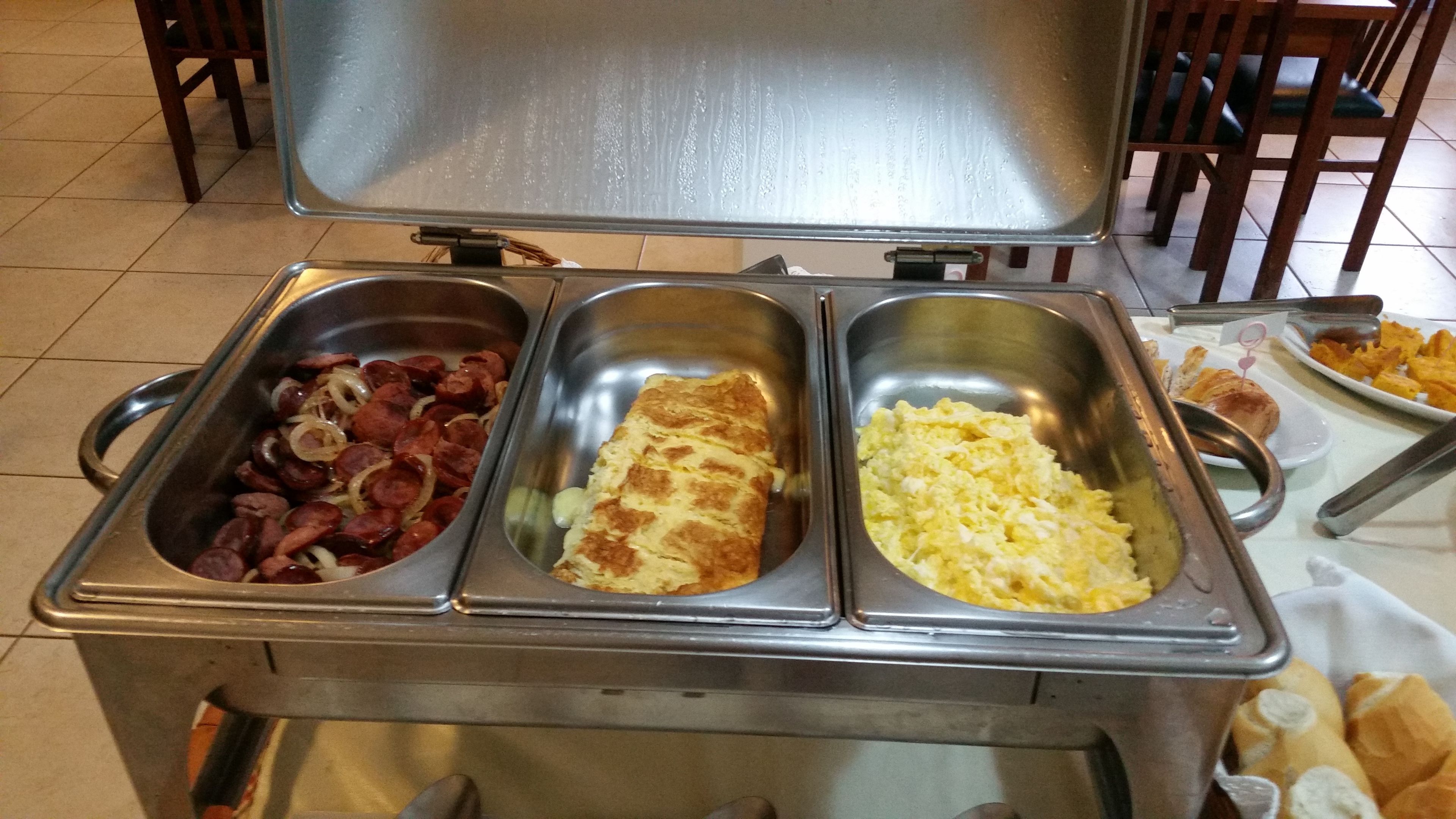 Un bufé con tres estaciones de comida: una con salchichas y cebollas, otra con huevos revueltos y otra con un plato horneado.