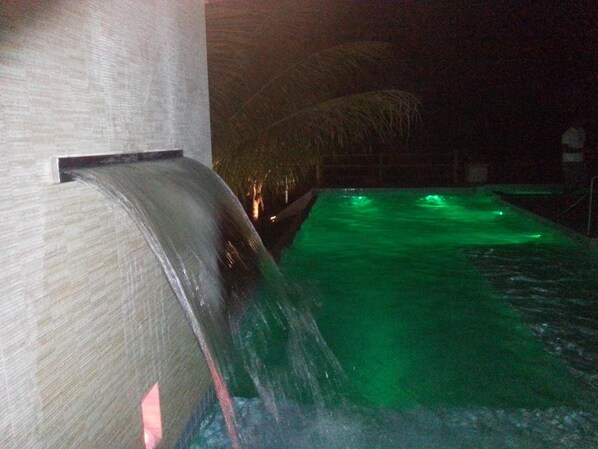 Une vue nocturne d’une piscine agrémentée d’une cascade et illuminée de lumières vertes.