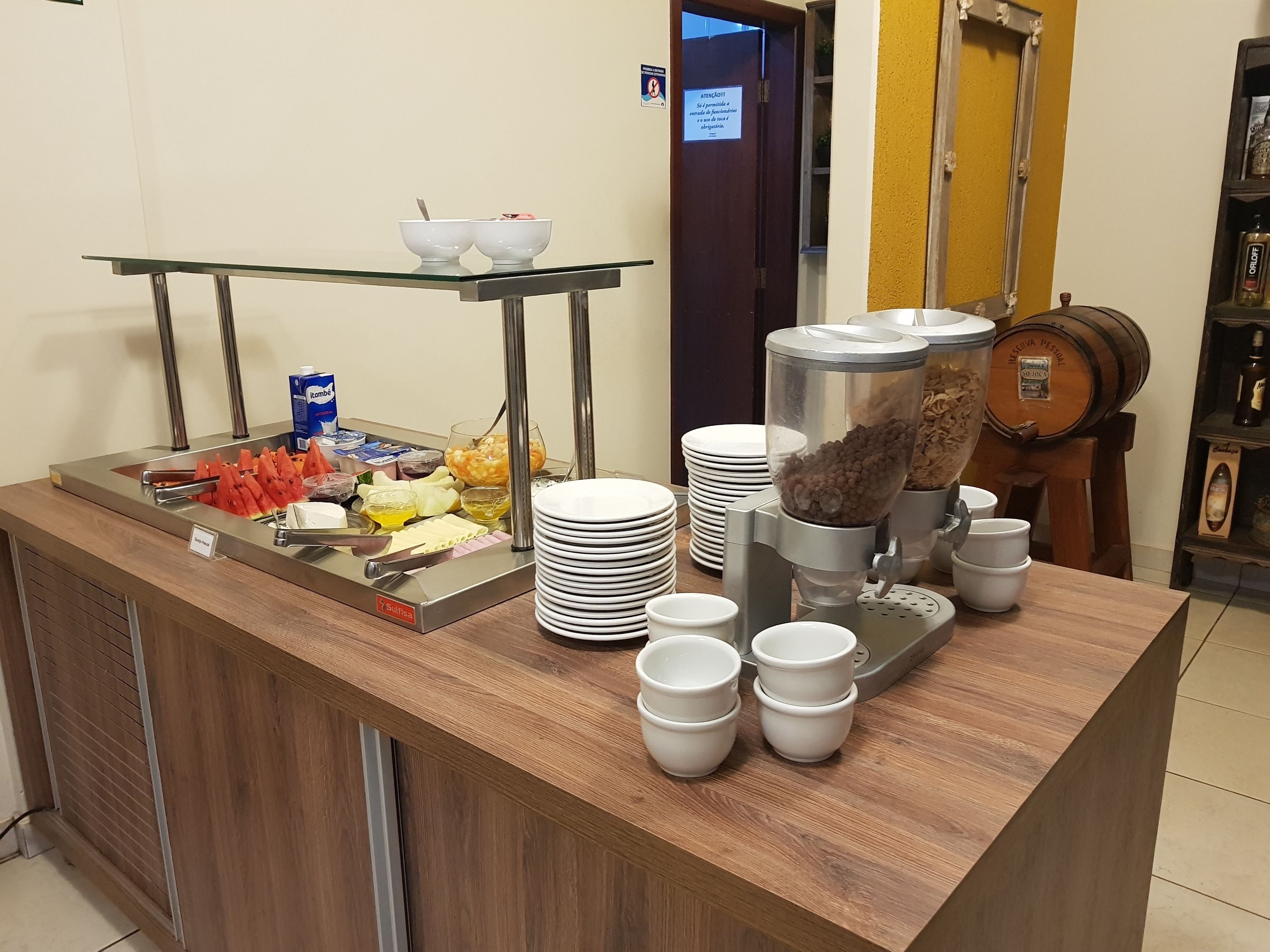 Montaje tipo bufé con diversas opciones de comida, como frutas, cereales y bebidas, dispuestas sobre una encimera de madera con un estante de vidrio arriba.