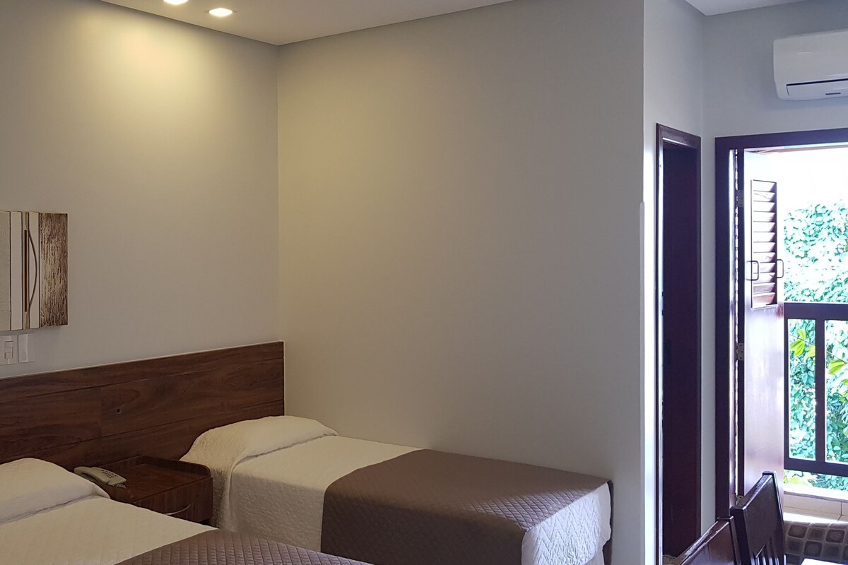 A hotel room with two beds, a wooden headboard, a wall-mounted artwork, and a window with a view of greenery.