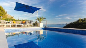 A clear blue swimming pool with a view of the sea, a lounge area with a sofa and umbrella, and a tiled deck. - Villa Levanda *** villa in Makarska, private pool (Makarska)