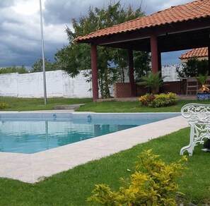 Una piscina con solárium de azulejos, un porche cubierto con pilares rojos y una pared blanca al fondo.