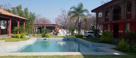 Una piscina con zona cubierta, rodeada de vegetación y un edificio con ventanas de arco.