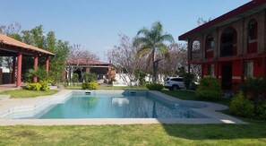 Una piscina con zona cubierta, rodeada de vegetación y un edificio con ventanas de arco.