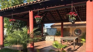 Un espace extérieur aménagé en patio, avec un sol carrelé, une table, des jardinières suspendues ornées de fleurs rouges et un pot contenant une plante aux fleurs jaunes.