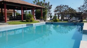 Ein Pool mit gefliestem Sonnendeck, umgeben von Grünflächen und einem überdachten Sitzbereich.