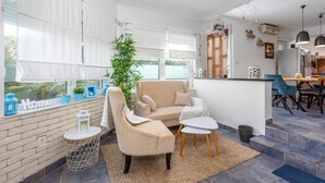A modern living room with a beige sofa, a small white table, and a potted plant. A kitchen area with a dining table and chairs is visible in the background. - Luxurious home in Kornic (Kornic)