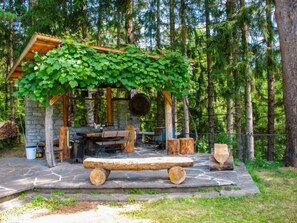 Een rustieke buitenzitplaats met een stenen tafel en banken, omringd door een stenen muur en een houten pergola begroeid met groene klimplanten.