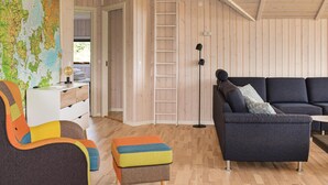 Una sala moderna con suelo de madera, un sofá gris oscuro, un sillón colorido y un pequeño otoman. Una escalera está apoyada en la pared, y se ve un mapa del mundo en la pared.