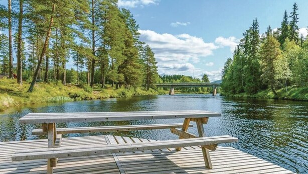 Een houten steiger met een picknicktafel met uitzicht op een rivier, omgeven door bomen en een brug in de verte.