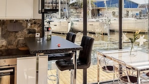 Una cocina moderna con encimera de mármol, comedor con mesa y sillas de vidrio, y vista a barcos a través de grandes ventanales.