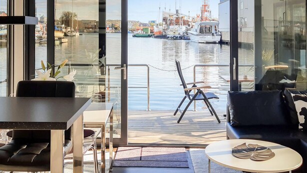 Un interior moderno con vistas a un puerto deportivo. Incluye un comedor con mesa y sillas, una zona de estar con sofá y una pequeña mesa redonda con un objeto decorativo.