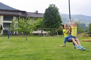 A grassy outdoor area with a swing set, a building with a balcony, and a mountainous backdrop. - Family Room - Ferienhof Stadlpoint (Ried im Zillertal)
